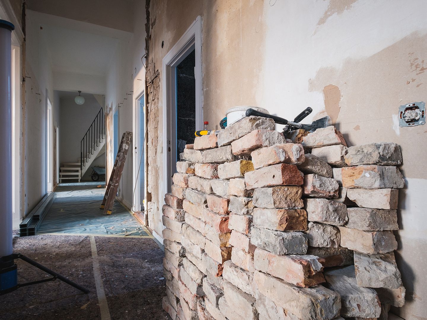Renovierungsszene mit einer aufgeschichteten Steinmauer, unfertigem Trockenbau, Werkzeugen und einer Treppe in einem gut beleuchteten Flur.