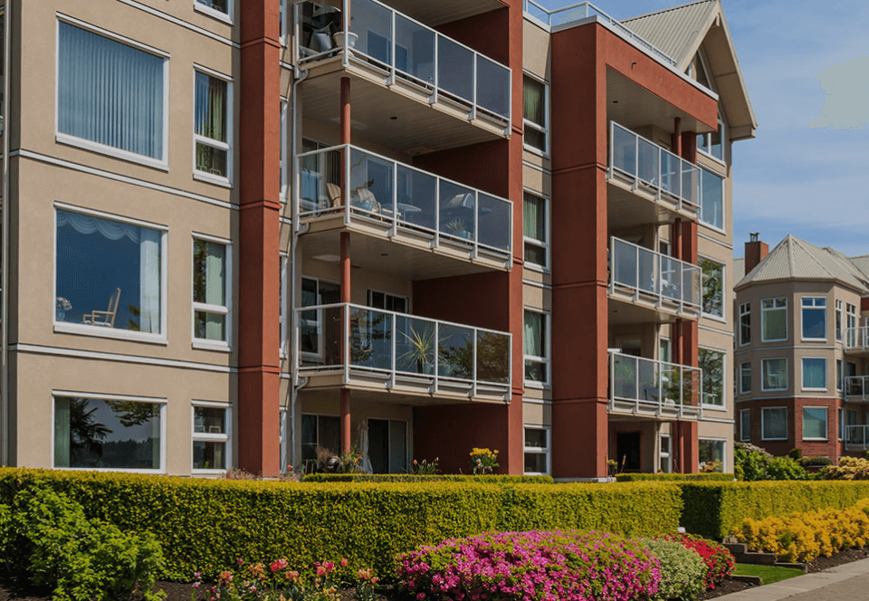 Modernes Wohnhaus mit Glasbalkonen, bunten Blumenbeeten und einem klaren blauen Himmel im Hintergrund.