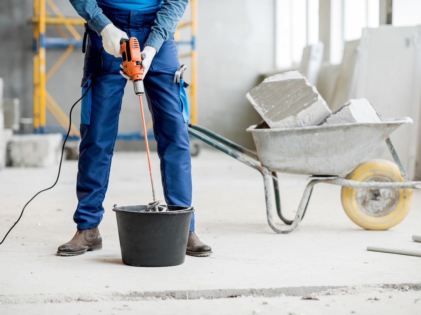 Ein Arbeiter in blauer Kleidung bedient auf einer Baustelle einen Mischer, während in der Nähe eine mit Material gefüllte Schubkarre steht.